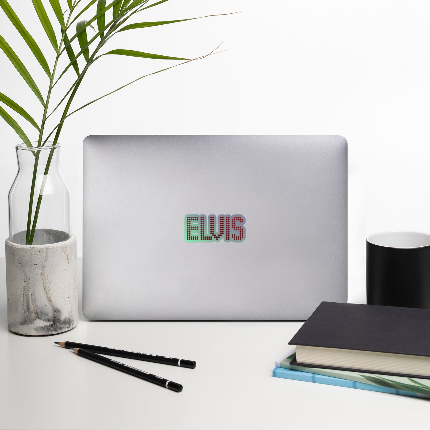 A silver laptop on a white desk with a black script decal reading 'ELVIS' on the back. The scene is styled with a glass vase holding green palm leaves, two black pencils, stacked notebooks, and a black cup against a clean white background.