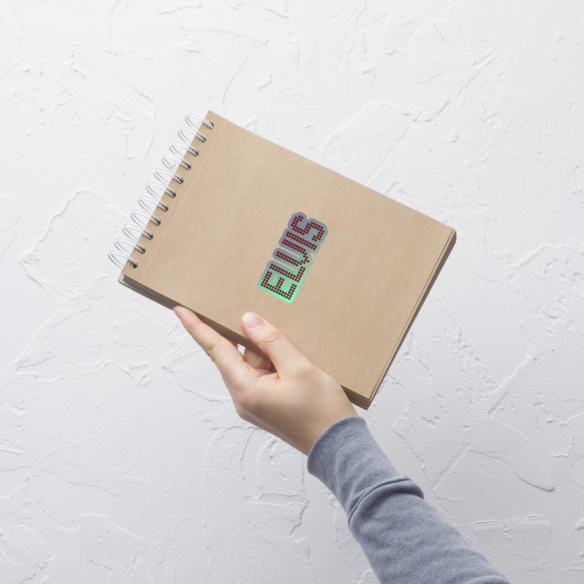 A hand holding a brown kraft paper spiral notebook with the name 'ELVIS' printed on the cover in a colorful, glittery font, set against a textured white wall.